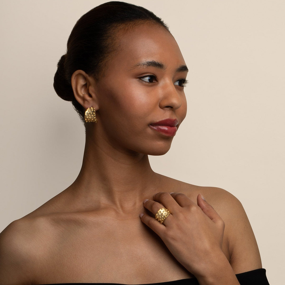 Woman wearing gold earrings and ring against a beige background
