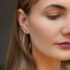 A close-up of a woman wearing large gold hoop earrings and a delicate gold necklace, with her face partially cropped and her hair visible against a soft gray background.