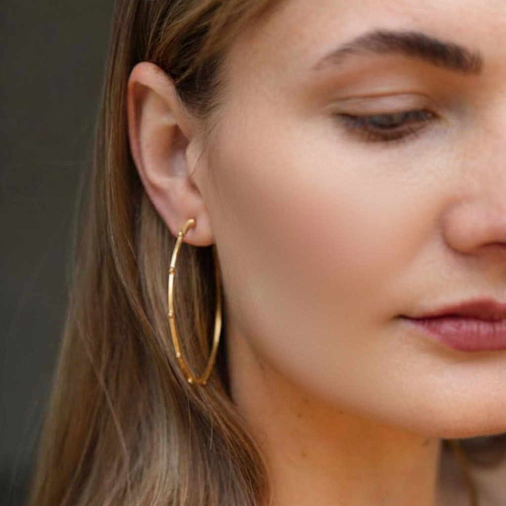 A close-up of a woman wearing large gold hoop earrings and a delicate gold necklace, with her face partially cropped and her hair visible against a soft gray background.